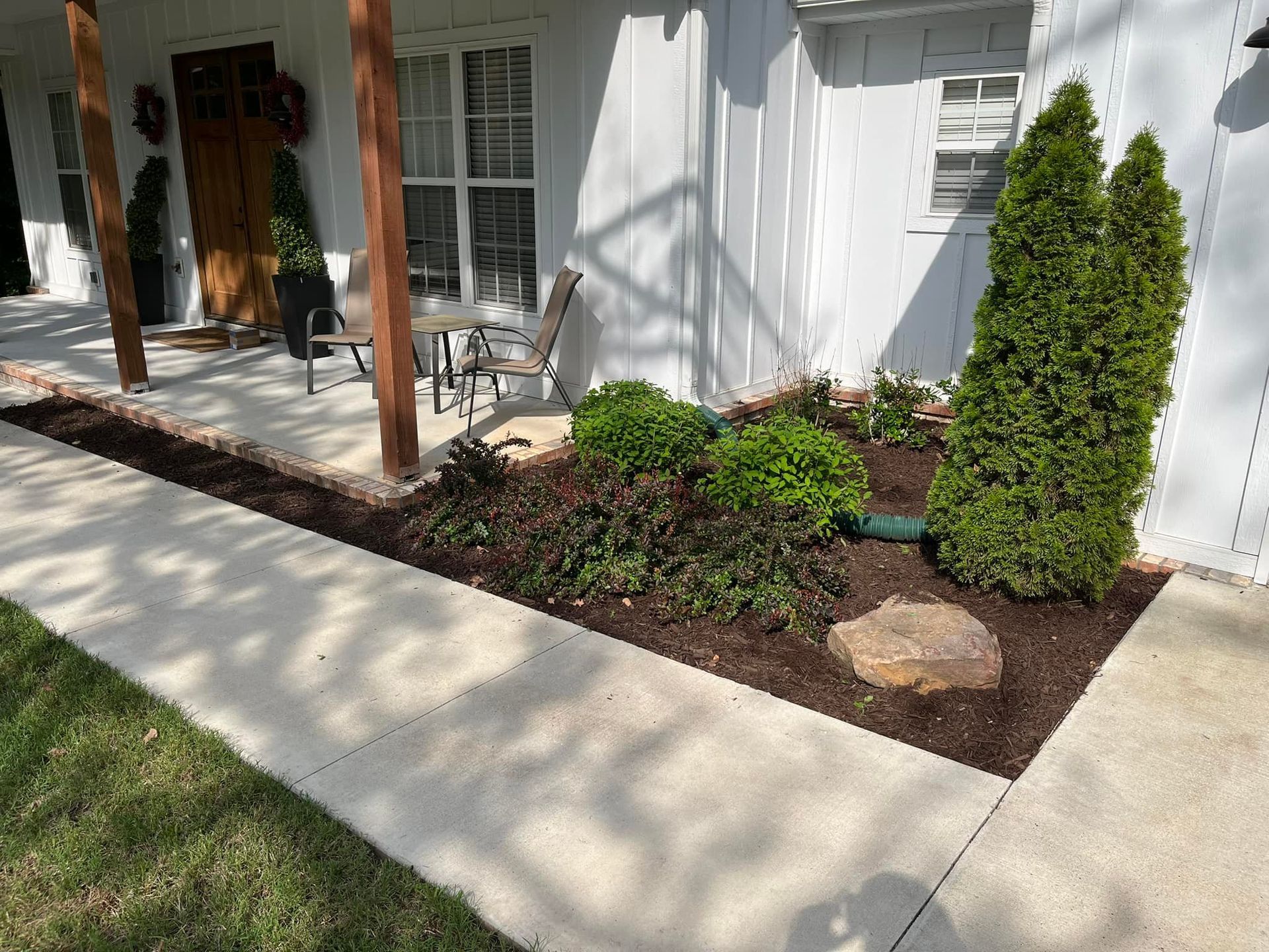 A landscaped flower bed beside a white house with a concrete walkway and lawn.