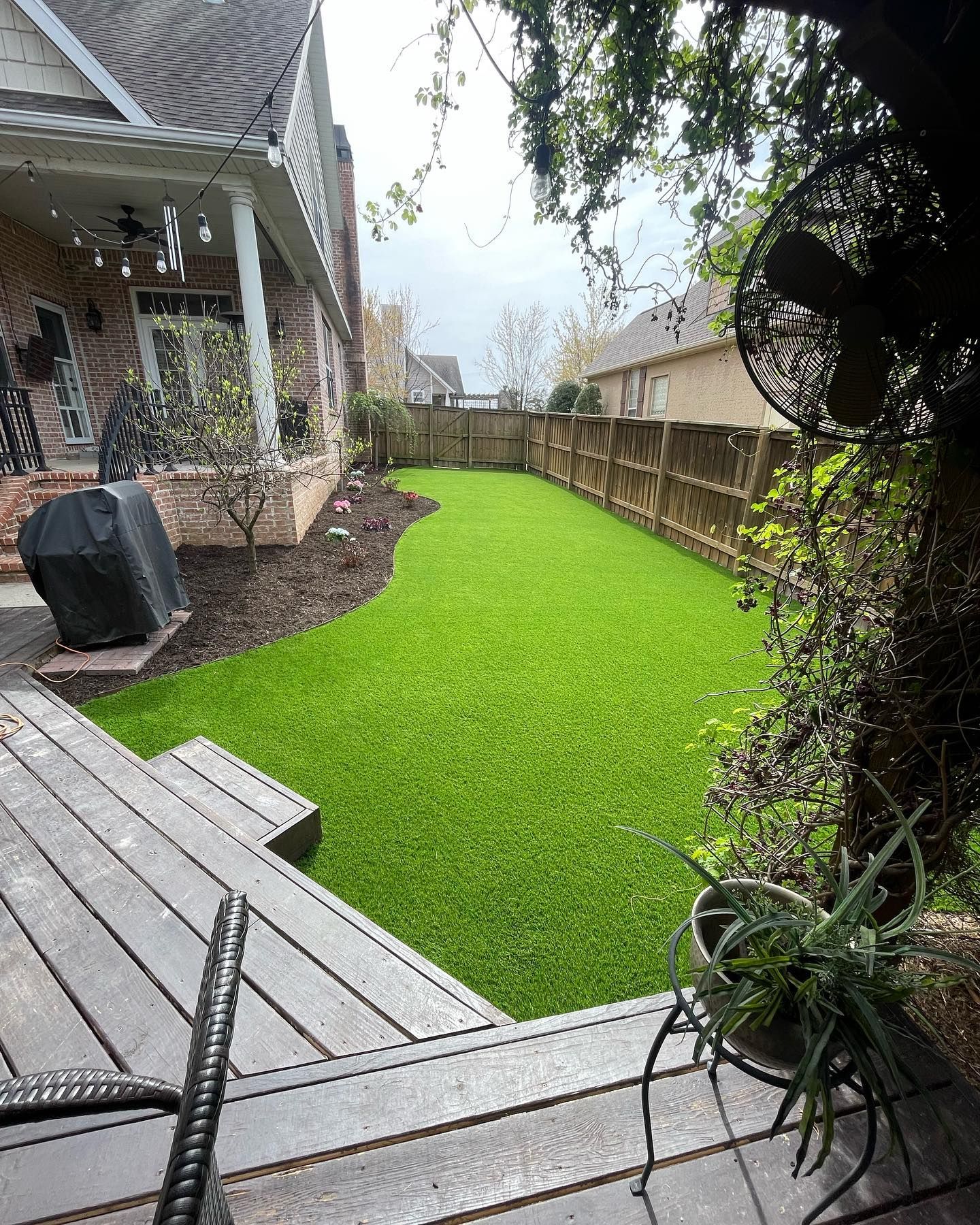 Backyard with green artificial turf, wooden deck, and a brick house.