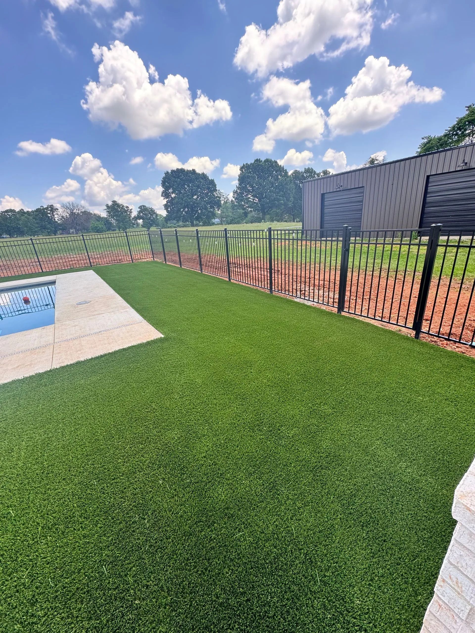 Green artificial turf yard next to a pool and black metal fence. Blue sky with puffy clouds