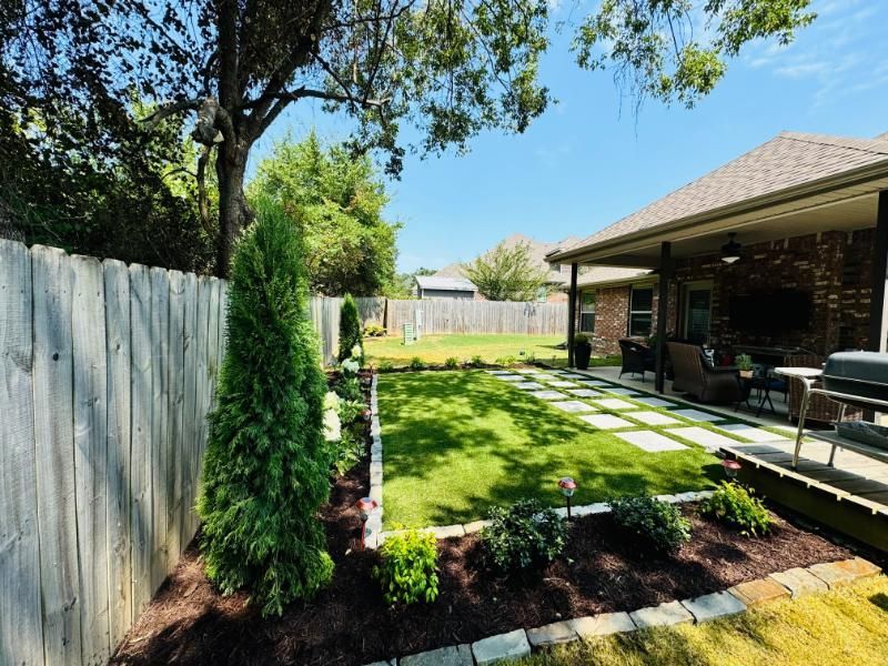 Well-manicured backyard with a patio, green lawn, garden beds, and a wooden fence on a sunny day.