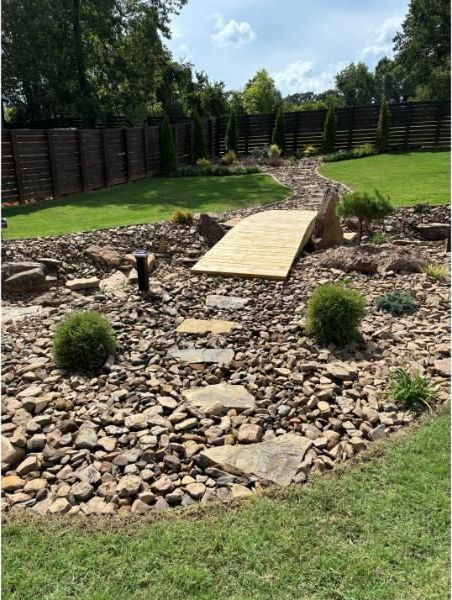A backyard garden with a wooden bridge over a rocky bed. Green lawn and trees surround it.