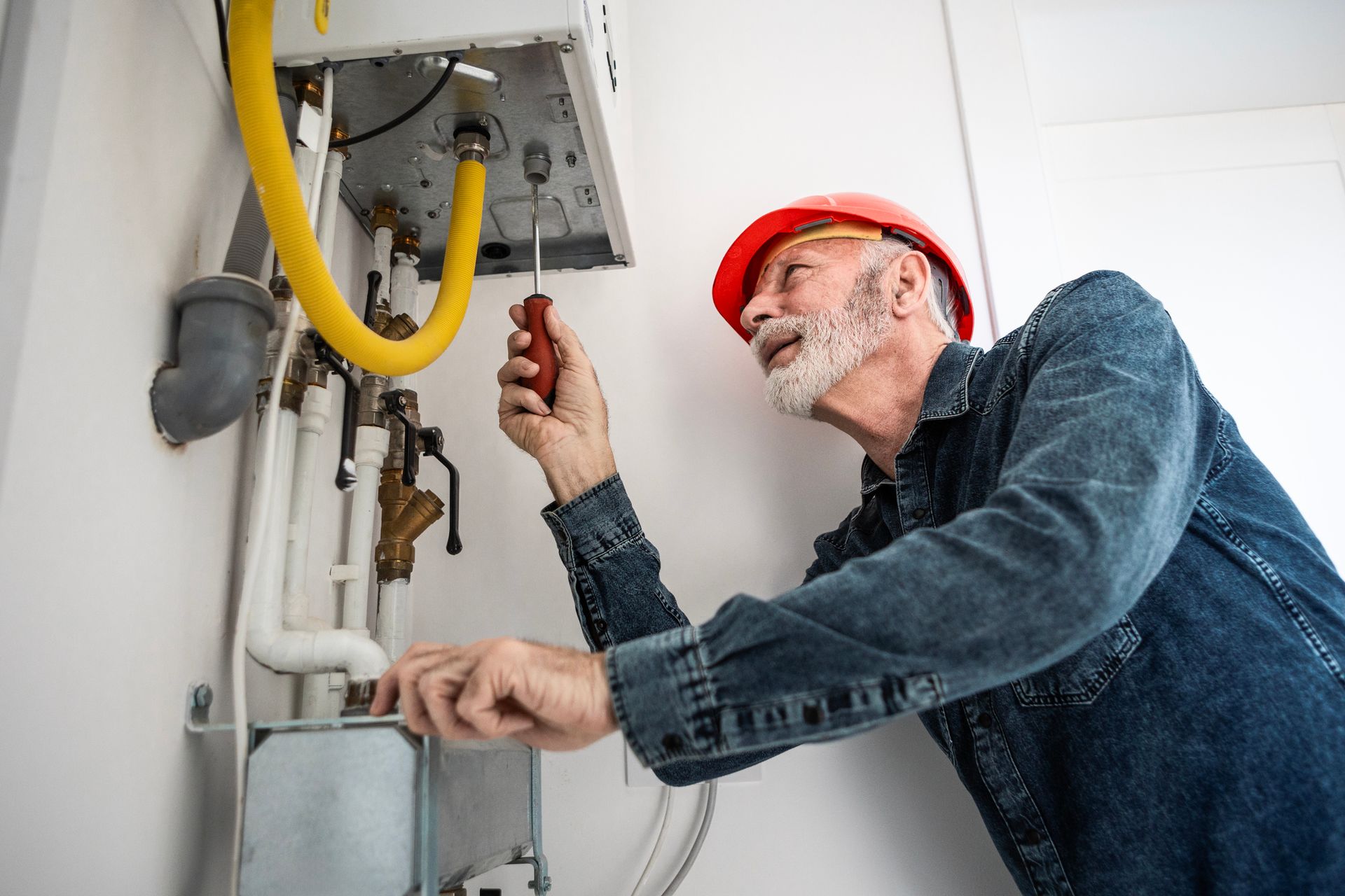 Senior technician in jean jacket and red hat expertly installing a home heating system.