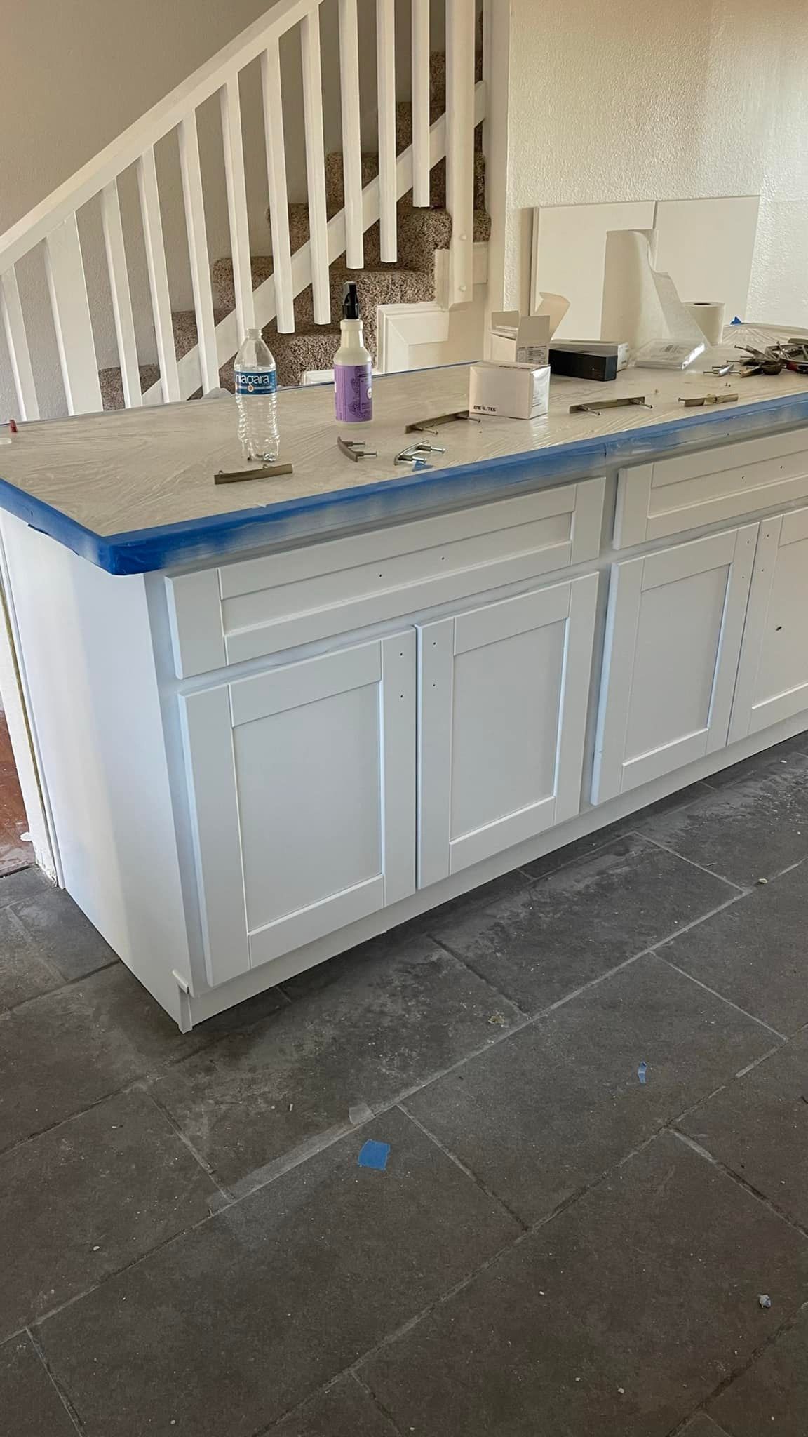 White kitchen island with cabinets, next to a staircase. Blue painter's tape, countertop and gray floor.