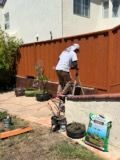 Person shoveling dirt into a planter by a brown fence. Outdoors, sunny day, bags of soil, and potted plants.