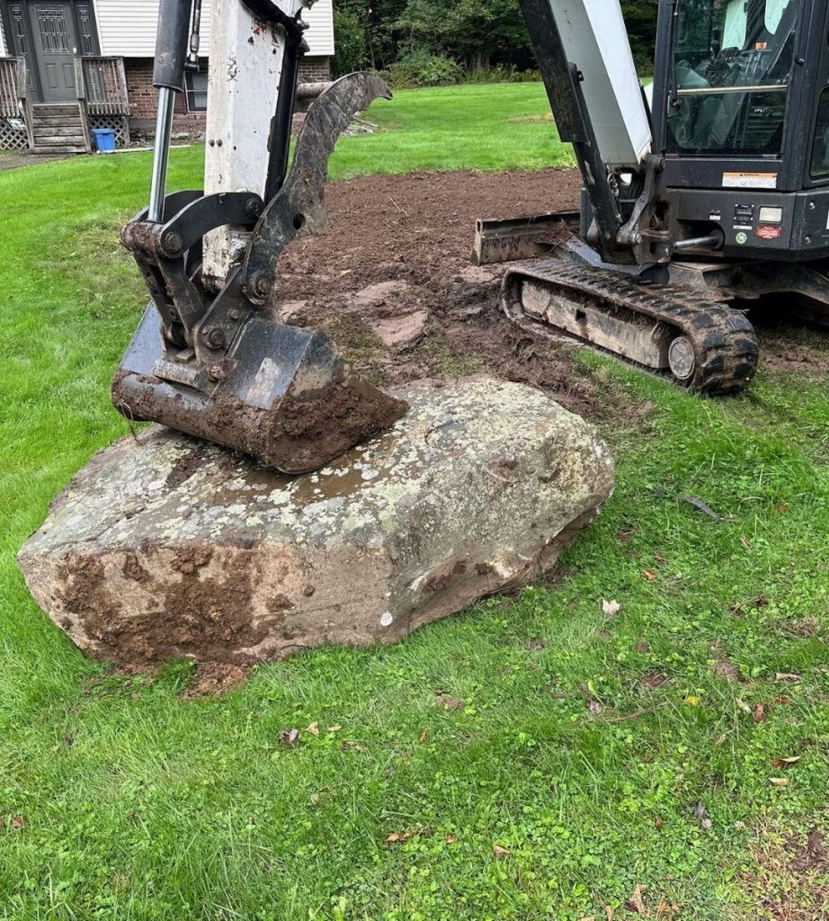 A bulldozer is digging a hole in the grass next to a large rock.