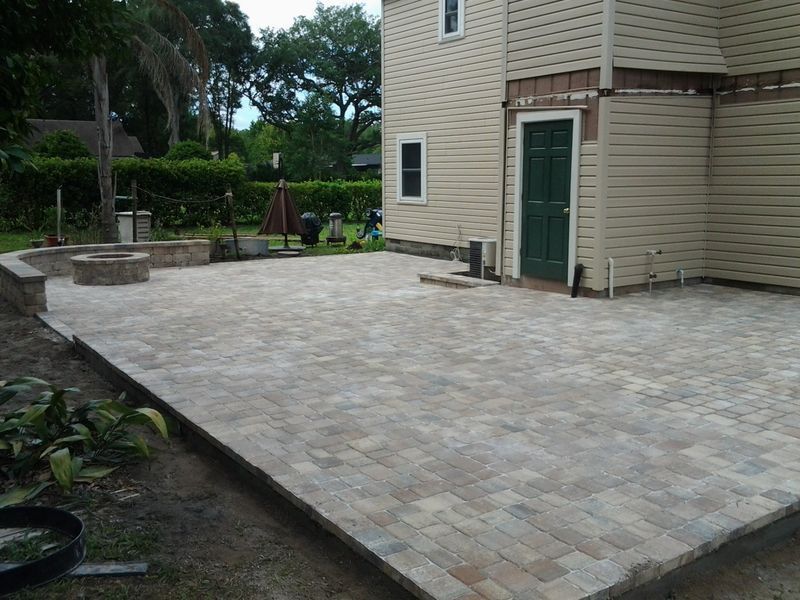 Brick paver patio with fire pit next to a two-story beige house, green door.