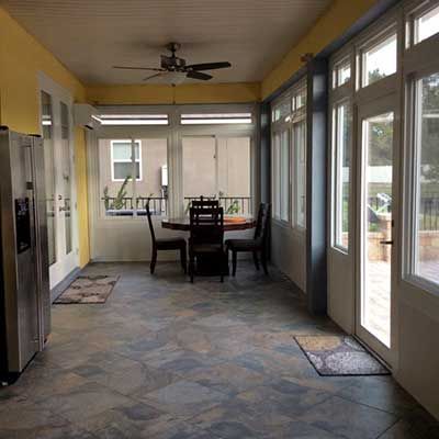 Sunroom with table, chairs, windows, and slate tile floor. Refrigerator on the left, rugs scattered.