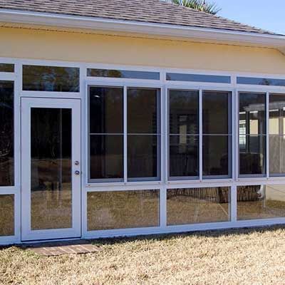 White-framed sunroom with glass windows and door attached to a yellow house, on a sunny day.
