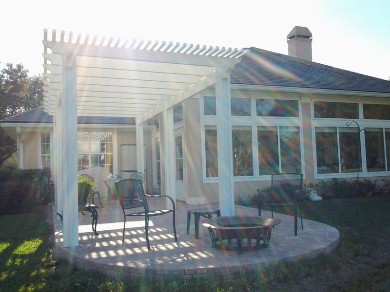 White pergola over a patio with chairs and fire pit near a house with large windows.