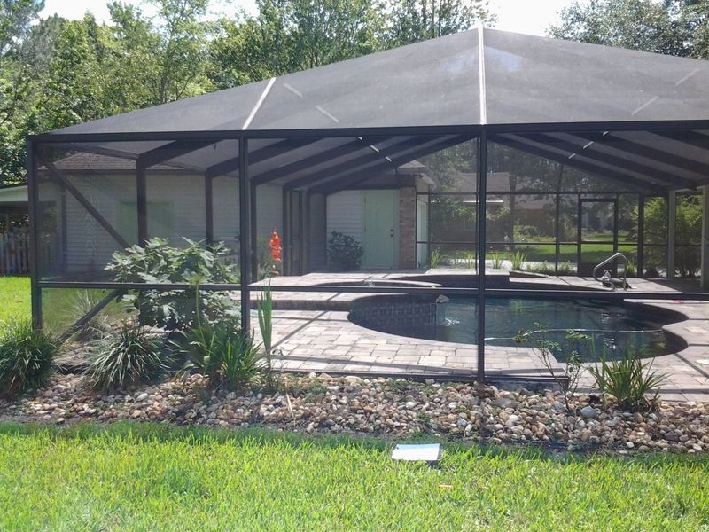 Screened-in pool enclosure with dark framing, a black roof, and a dark-bottomed pool. Set on a patio with landscaping.