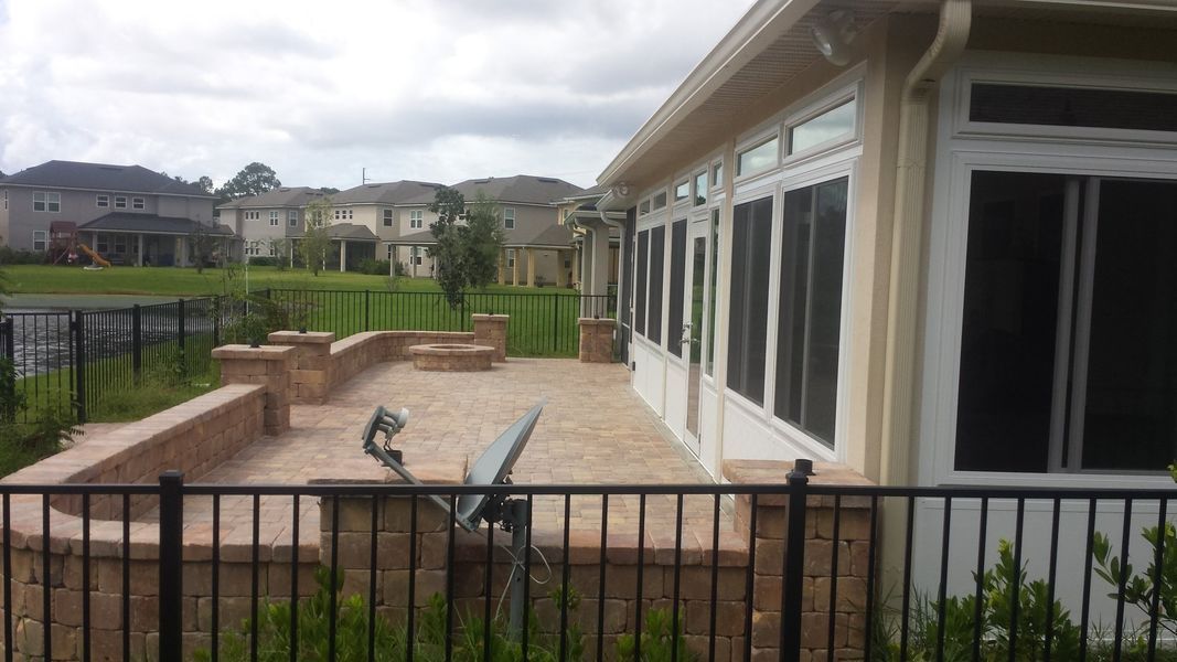 Patio with brickwork, black fence, and building with screened windows. Houses and green lawn in background.