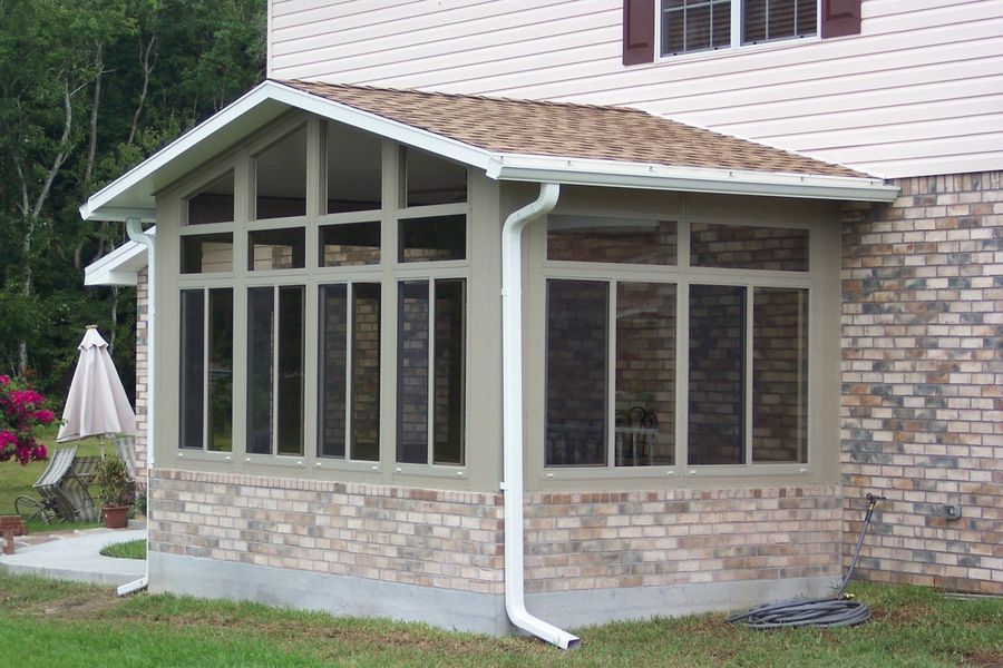 Tan brick and glass sunroom addition to a brick-sided house, with a brown shingled roof and white gutters.