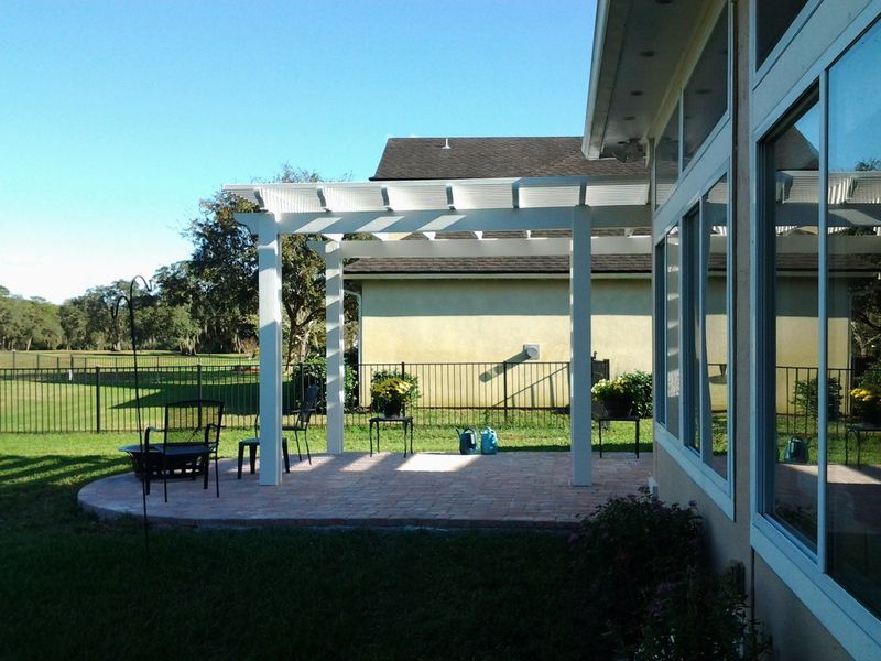 White pergola over a patio next to a house with large windows, overlooking a green lawn and golf course.