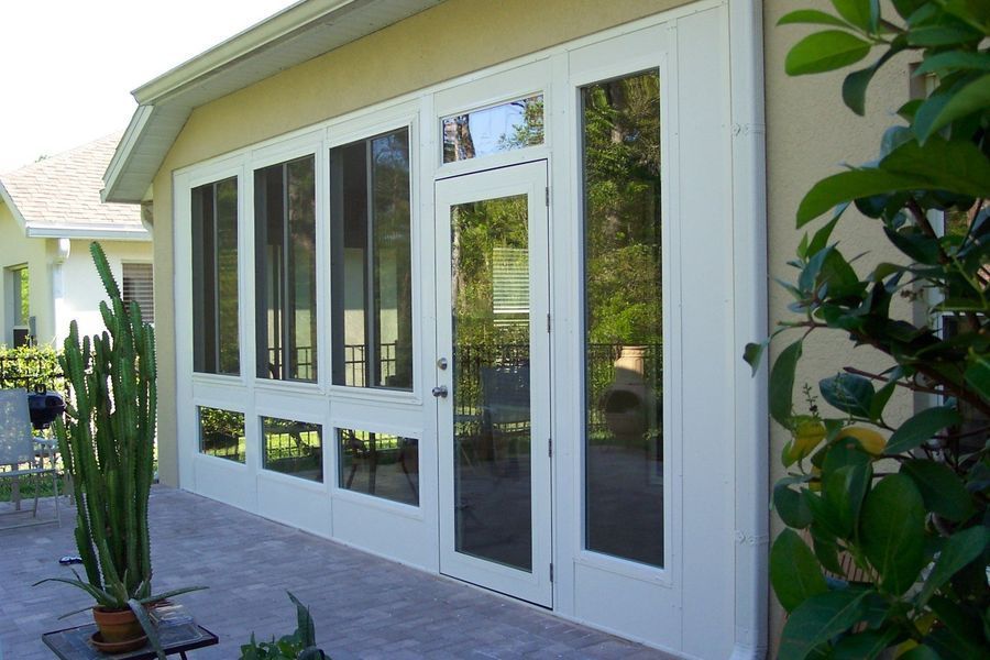 White-framed sunroom with glass windows and door attached to a light-colored building, on a patio with plants.