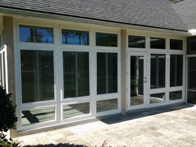 White-framed sunroom with multiple glass windows and doors; exterior view with brick patio and dark roof.