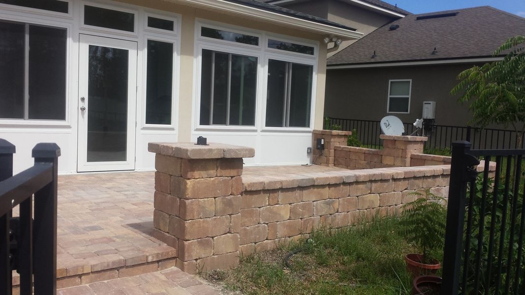 Patio with brick retaining wall, glass doors and windows, black railing, and brown tile flooring.