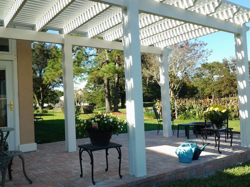 White pergola over a brick patio with wrought iron tables and potted flowers, overlooking a green lawn.