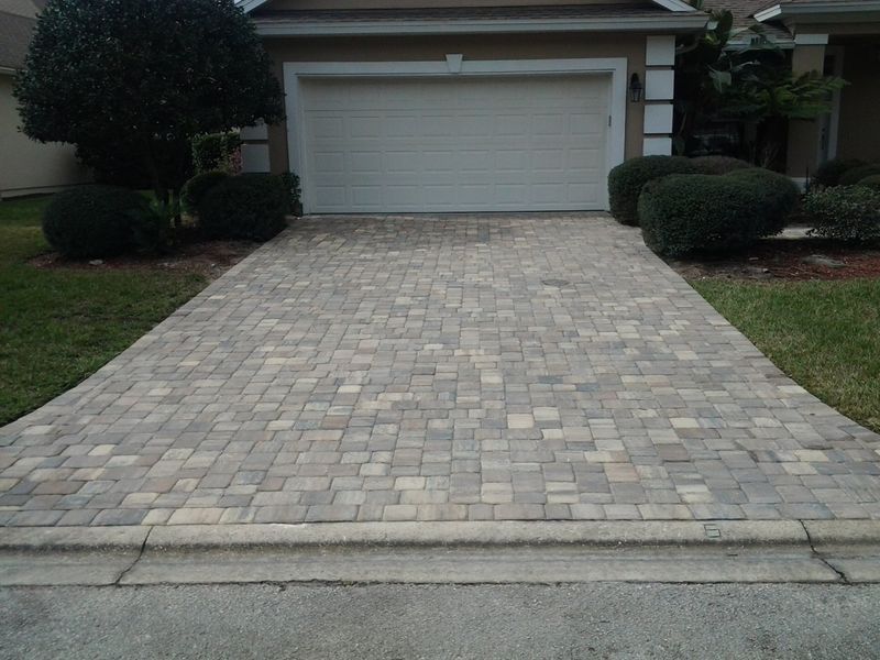 Brick driveway leading to a garage.  Driveway is light gray, edged with a curb.  Green bushes and grass on either side.