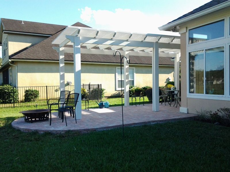 White pergola over a brick patio with outdoor furniture, next to a beige house, on a sunny day.