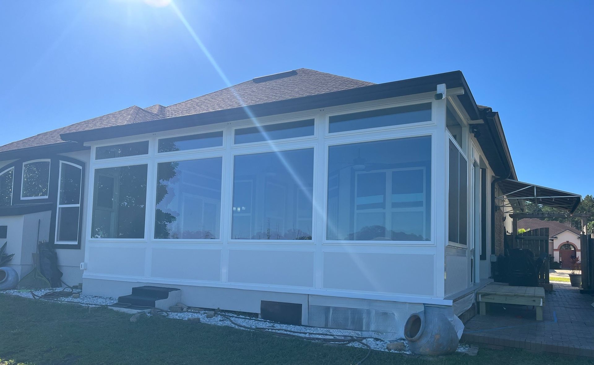 A sunroom with large windows, white trim, and a dark roof. Situated next to a house under a clear, blue sky.