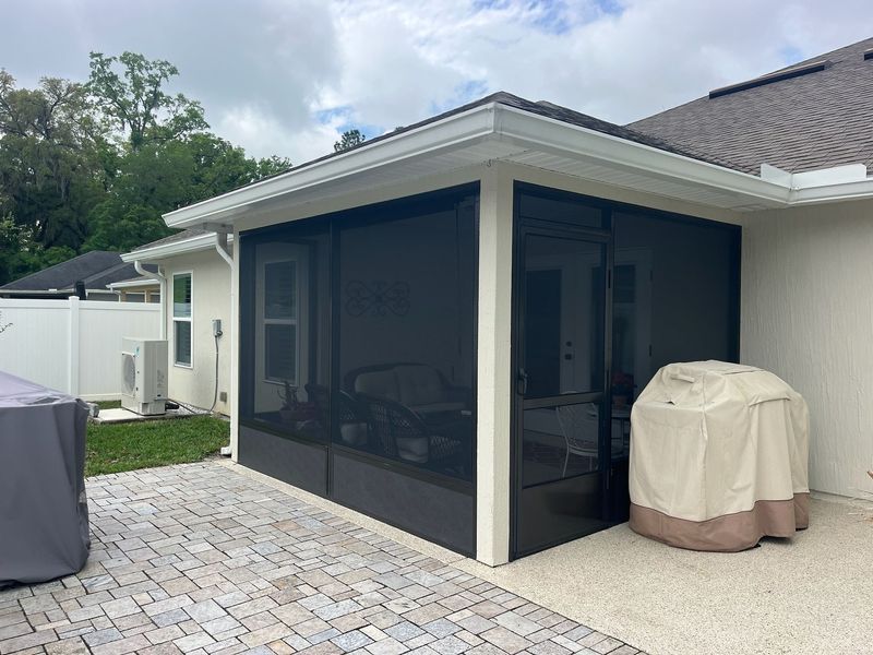 Screened-in patio with dark screens and beige grill cover on a brick and concrete patio next to a house.