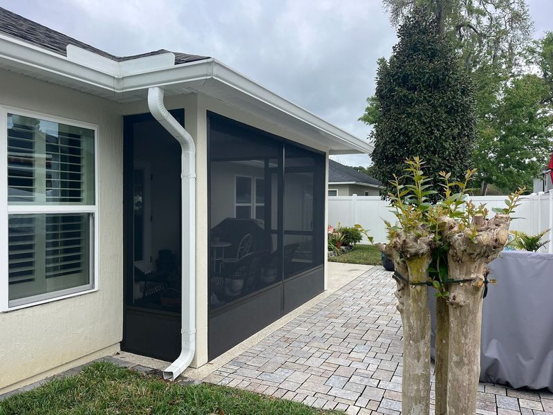Screened porch attached to a light-colored house. Dark screen, white trim, and a brick patio.