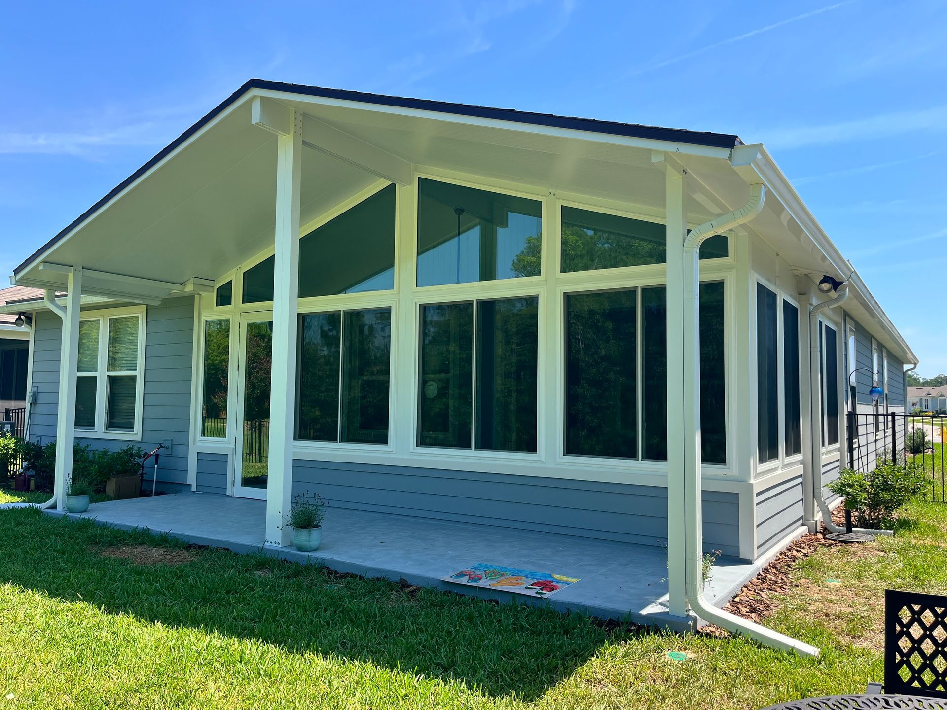 Grey sunroom addition with white trim, glass windows, and a concrete patio on a sunny day.
