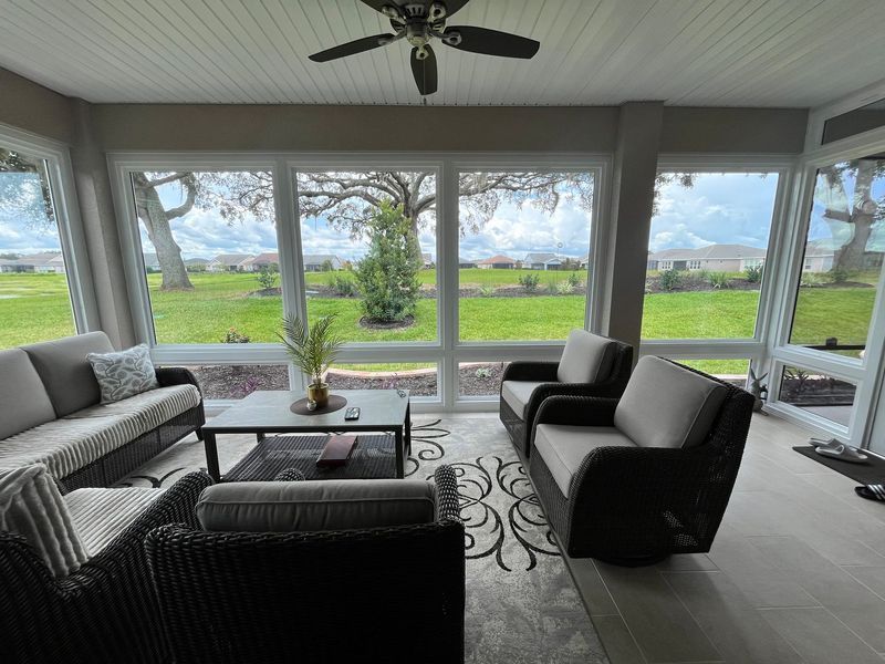 Sunroom with sofas, coffee table, and large windows overlooking a green lawn under a cloudy sky.