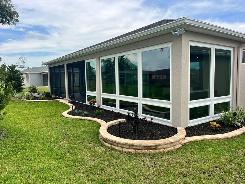 A beige sunroom with large windows, dark trim, and a low stone wall planter in a grassy yard.