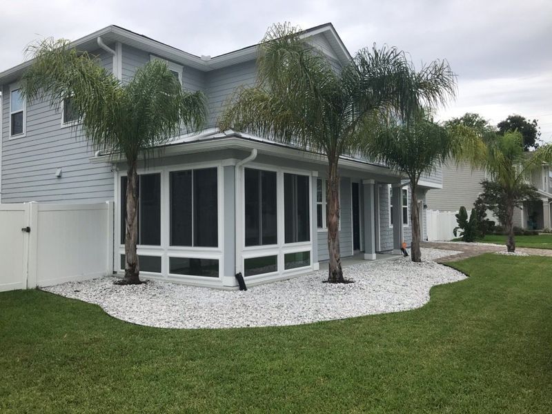 Backyard with screened-in porch, light gray house, palm trees, white gravel, and green grass.