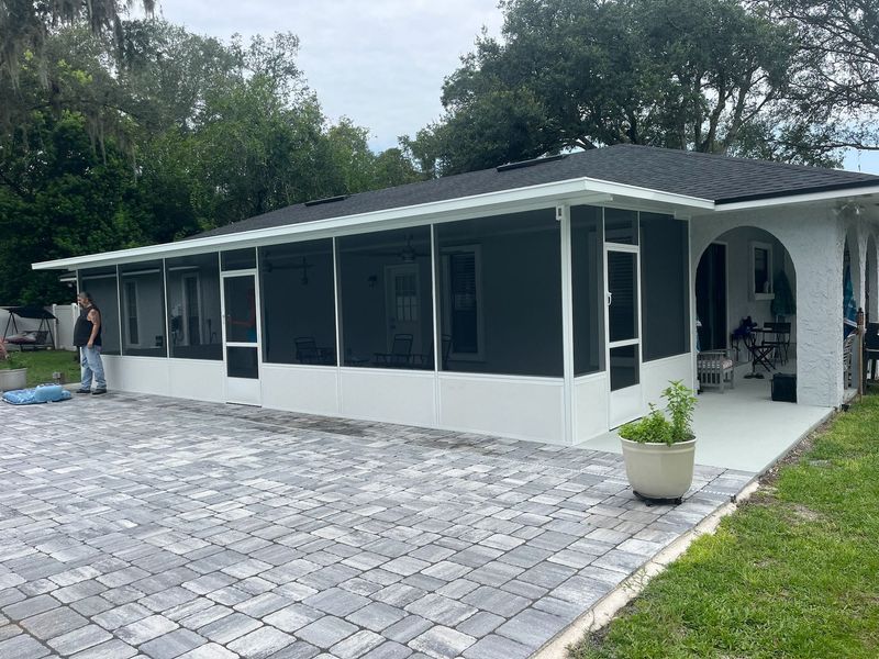 Screened-in porch addition with a paver patio. White trim and dark screens. Person stands near the porch.