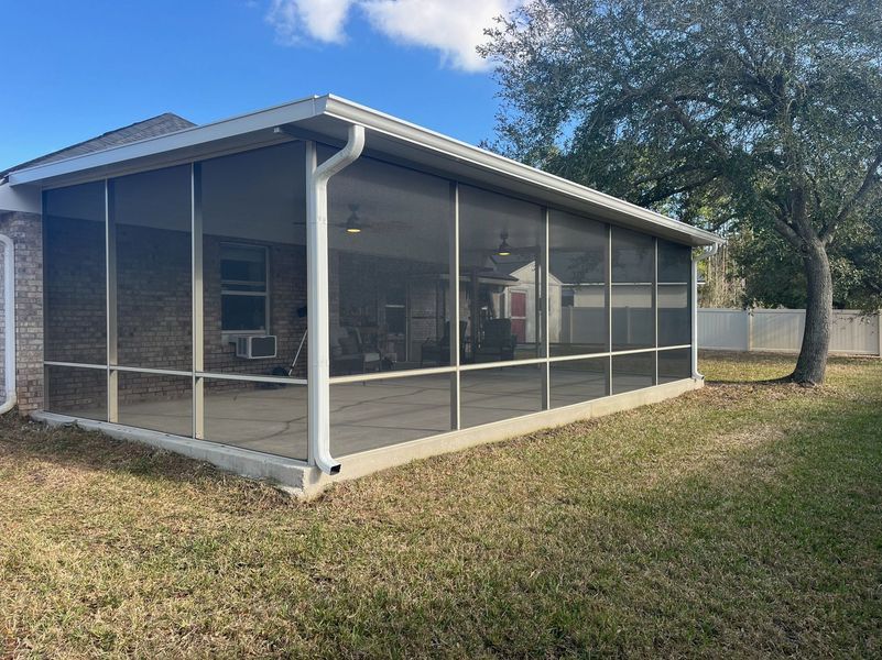 Screened-in patio extension attached to a brick house. Concrete flooring, white trim, and a tree nearby.
