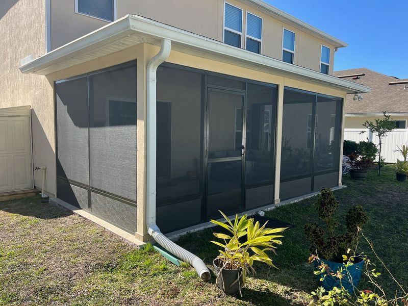 Screened-in porch attached to a light-colored house. Black screens, beige trim, green grass, and small plants.