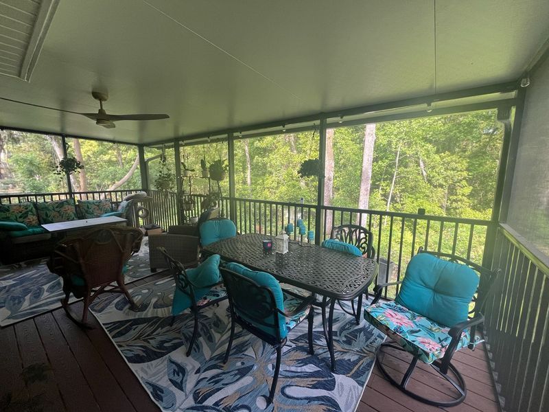 Screened-in porch with outdoor furniture, overlooking a green wooded area. Teal cushions and decor.
