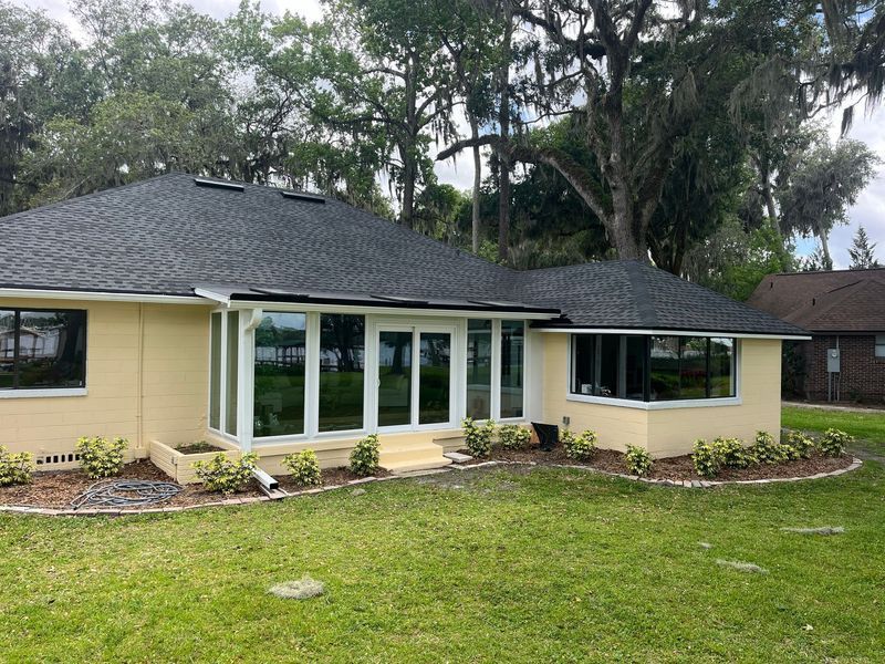 Yellow house with black roof, large windows, and manicured landscaping in a grassy yard.