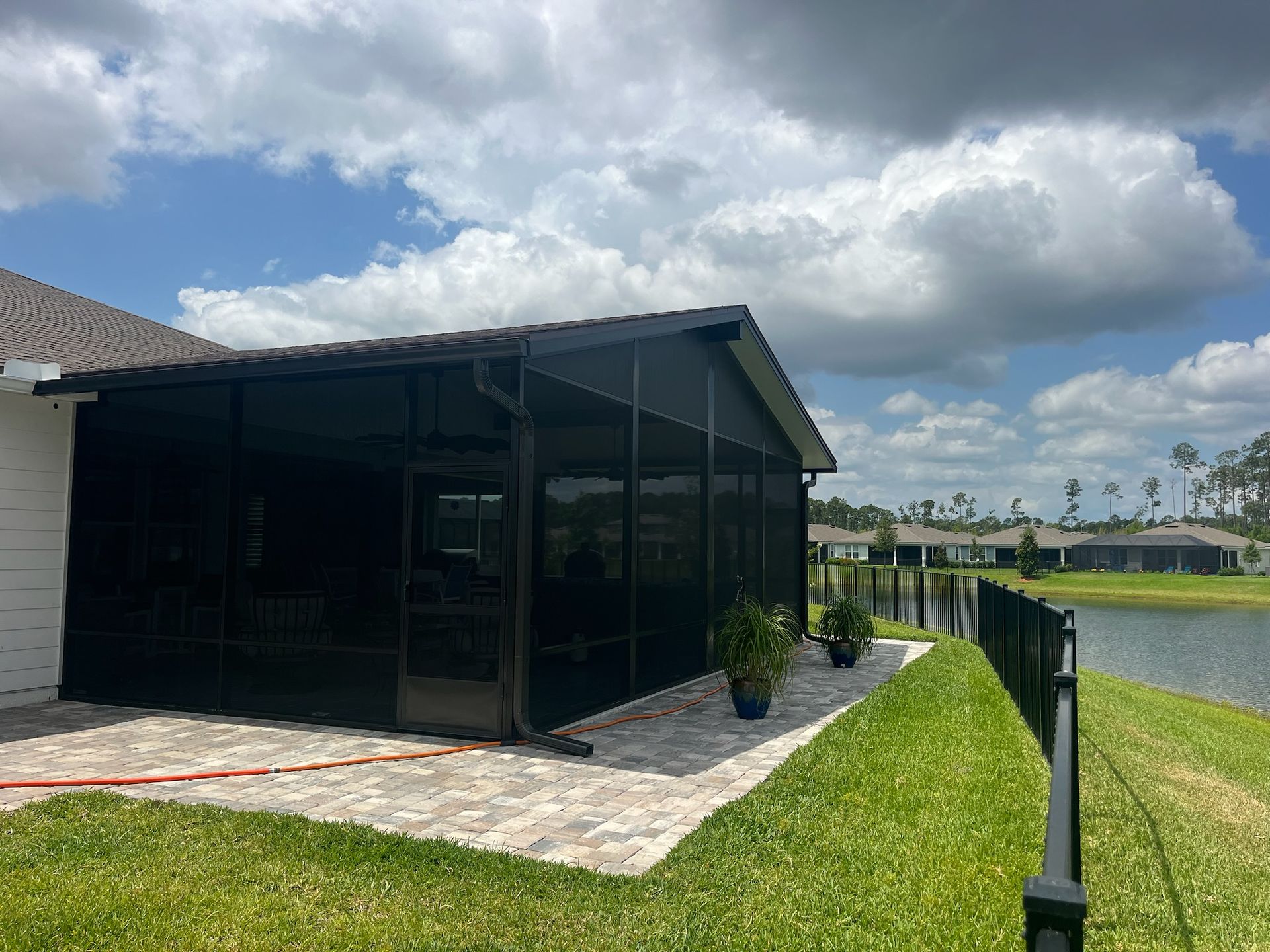Screened porch with dark screens, brick patio, and black fence next to a lake.