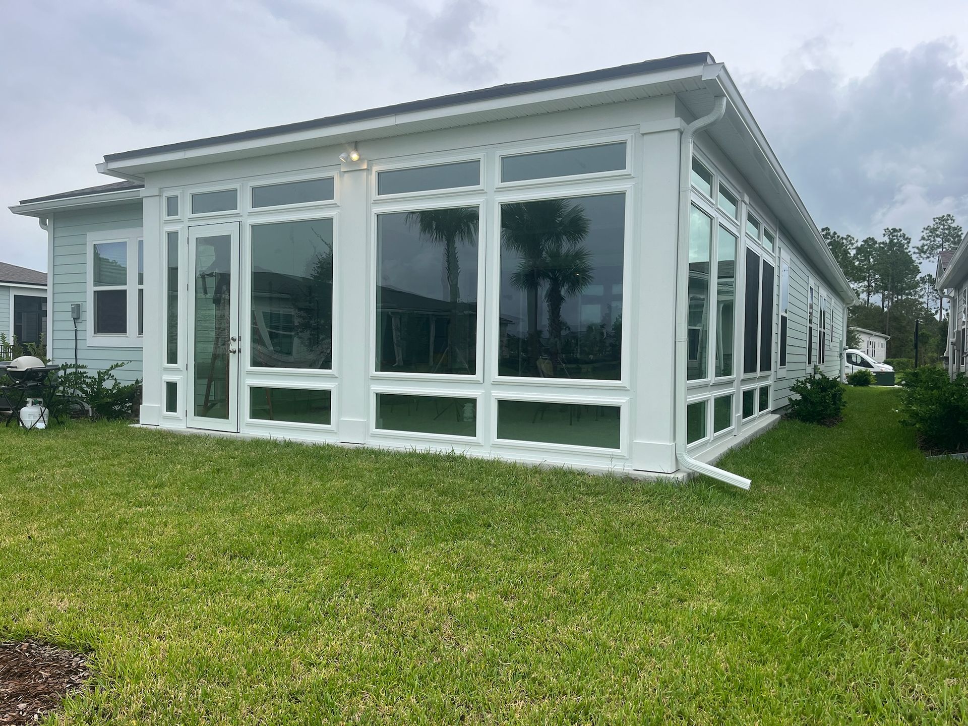 White sunroom with large windows, set on a green lawn, next to a light blue house, cloudy sky.
