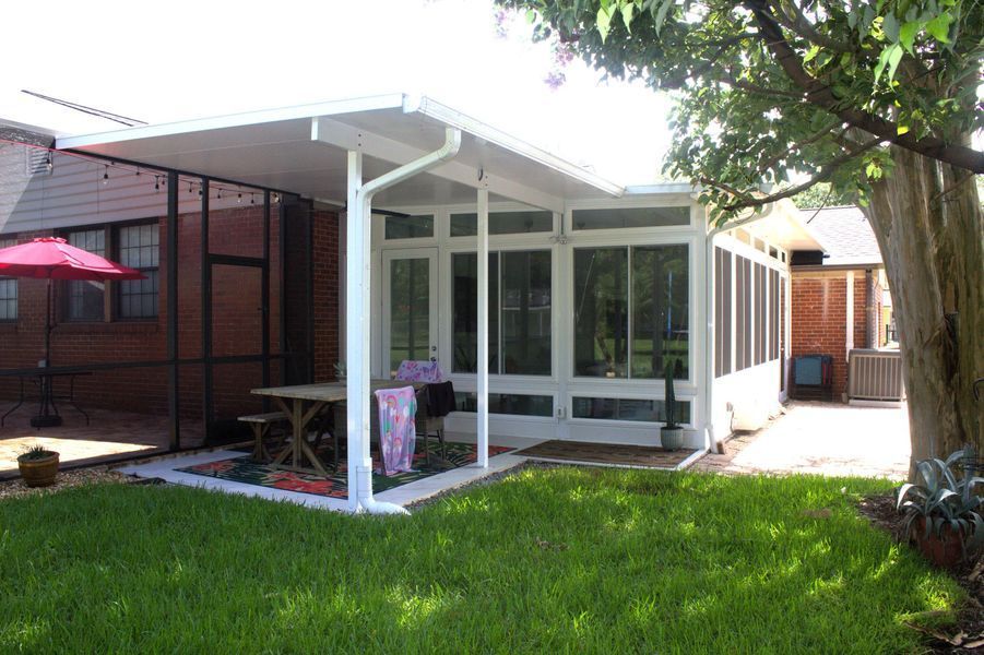 White-framed screened-in porch and patio with white roof attached to a brick home. A table sits on the patio.