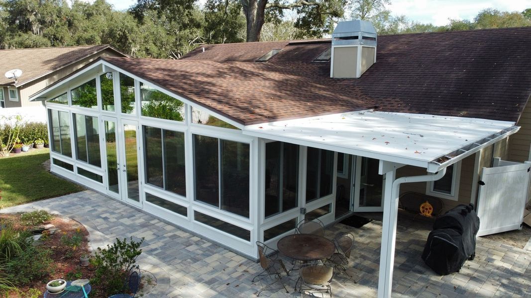 White-framed sunroom and covered patio attached to a house with a brown roof and chimney, on a paved area.