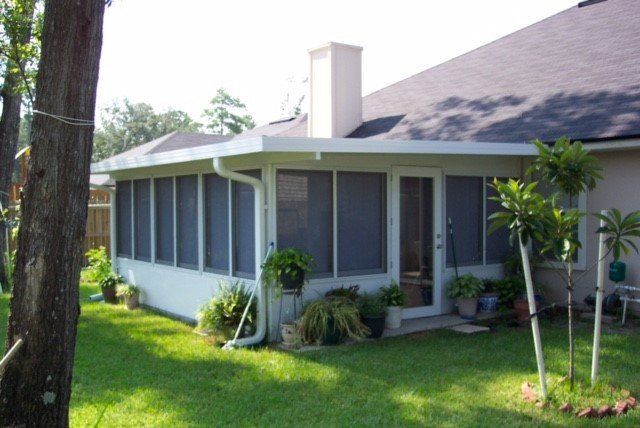 Screened-in porch attached to a house with plants, a lawn, and trees on a sunny day.