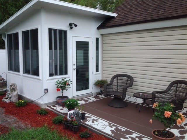 White sunroom addition with brown chairs, potted plants, and red mulch alongside a house with tan siding.