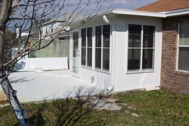 White sunroom attached to a brick building; glass windows, door, and a light-colored patio.