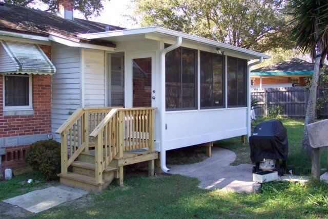 House with screened porch, wooden steps, and a grill on a concrete path in a yard.