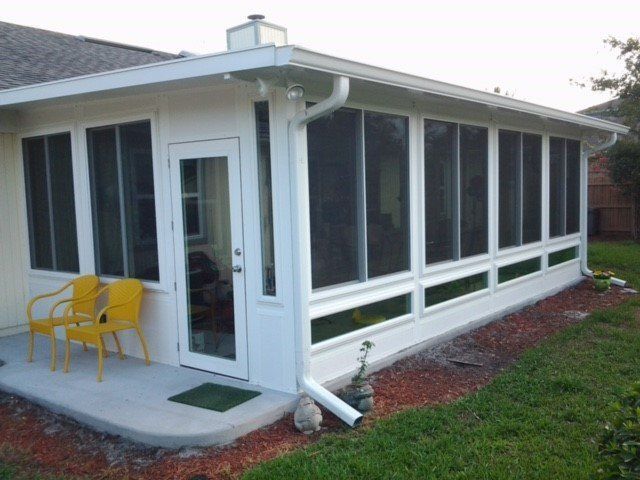 White sunroom with large screened windows, a door, and yellow chairs on a concrete patio, adjacent to a house.