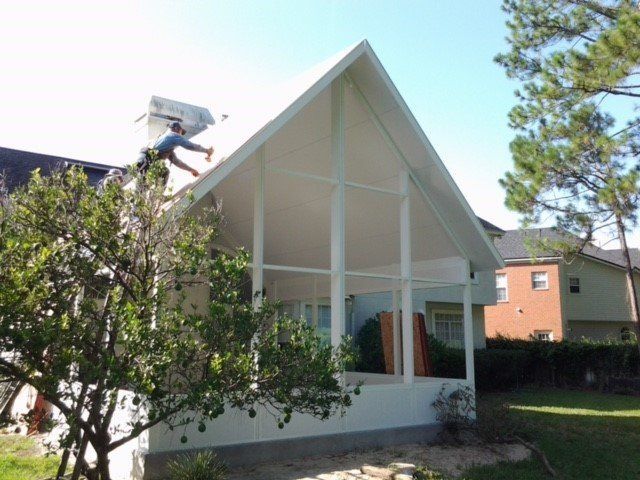 Man painting the roof of a white, open-air structure attached to a house; green trees and lawn surround it.