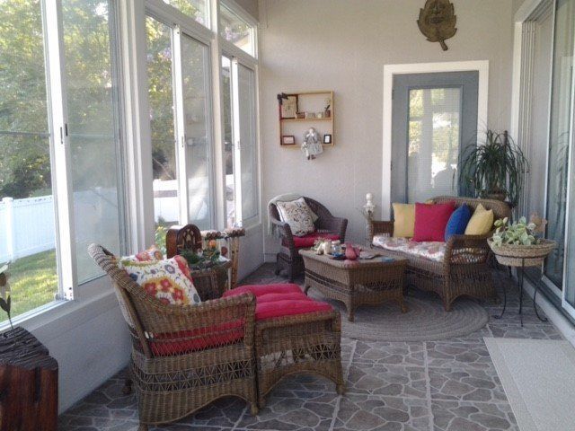 Sunroom with wicker furniture and colorful cushions. Beige walls, tiled floor, and large windows.