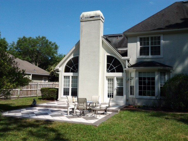Backyard patio with checkered tiles and outdoor furniture near a house with a tall chimney.
