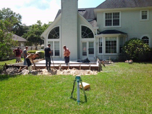 Construction workers pouring concrete for a patio in a backyard with a large house. Green grass and blue sky.