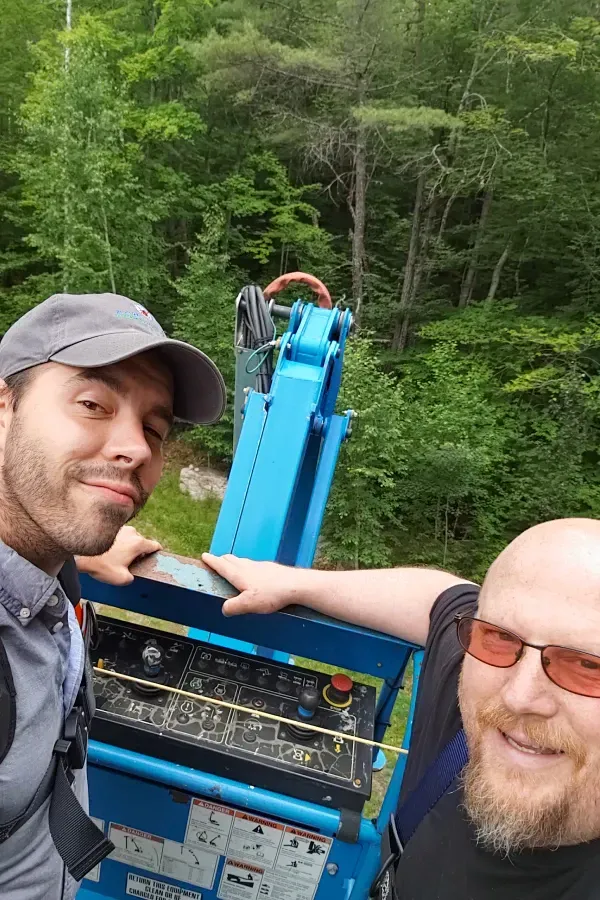Two men are posing for a picture in front of a crane in the woods.