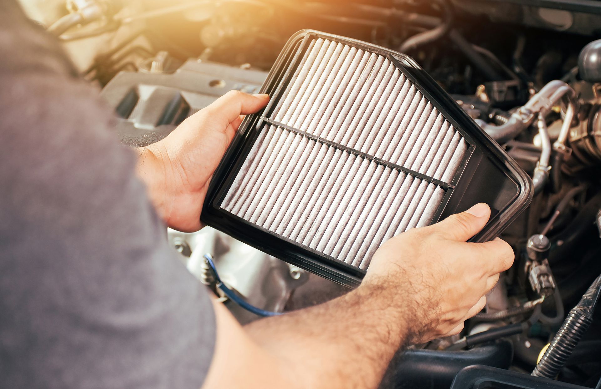 Hands holding a new car air filter, engine bay visible in the background.